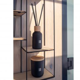 Dark gray scented candle with a wooden lid, placed on a metal shelf.