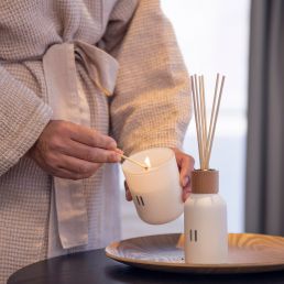 A white scented candle and diffuser on a brown tray, a person lighting the candle. The candle and diffuser bottle are decorated with minimalist patterns.